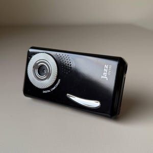 May include: A black digital camcorder with the text "Jazz DV150". The camcorder has a silver lens and a silver accent below the lens. The words "DIGITAL CAMCORDER" are printed on the front.