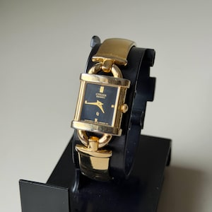May include: A gold-toned Citizen Quartz watch with a rectangular black face. The watch has gold hands and hour markers. The watch is displayed on a black stand.