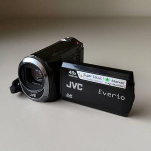 May include: A black JVC Everio camcorder with a flip-out screen. The screen displays text including 45x Dynamic Zoom, Super LoLux for increased brightness, and Advanced Image Stabiliser. The lens area features a silver ring and JVC branding.