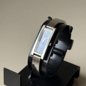 May include: A rectangular Citizen watch with a silver strap and a light blue face. The watch is displayed on a black stand. The watch face has a subtle iridescent sheen. The watch is a classic accessory.