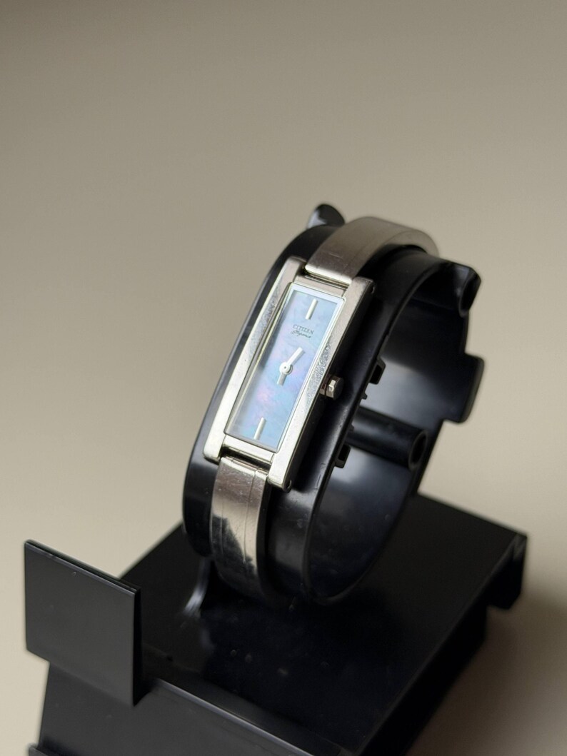 May include: A rectangular Citizen watch with a silver strap and a light blue face. The watch is displayed on a black stand. The watch face has a subtle iridescent sheen. The watch is a classic accessory.