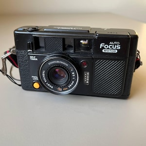 Puede incluir: Cámara de película Yashica Auto Focus Motor negra de 38 mm con correa roja y negra. La cámara tiene temporizador automático y bloqueo de enfoque. La lente es Yashica Lens 1:2.8. Sobre una superficie gris claro.