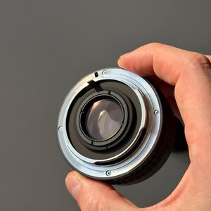 Op de afbeelding: Een close-up van een cameralens met een zwart en zilverkleurig metalen lichaam. De lens heeft een cirkelvormig ontwerp met meerdere schroeven en een reflecterend glaselement. De achtergrond is effen grijs.
