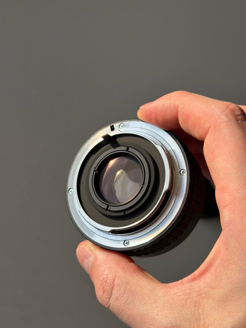 Op de afbeelding: Een close-up van een cameralens met een zwart en zilverkleurig metalen lichaam. De lens heeft een cirkelvormig ontwerp met meerdere schroeven en een reflecterend glaselement. De achtergrond is effen grijs.
