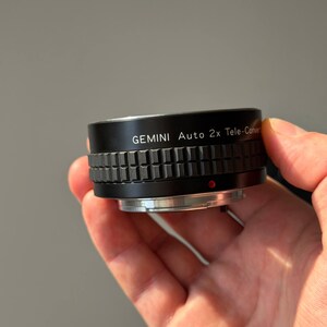 Op de afbeelding: Een zwarte Gemini Auto 2x Tele-Converter lens met een geribbelde grip. De lens heeft zilveren accenten en een rode stip. De tekst "GEMINI Auto 2x Tele-Converter" staat op de lens.