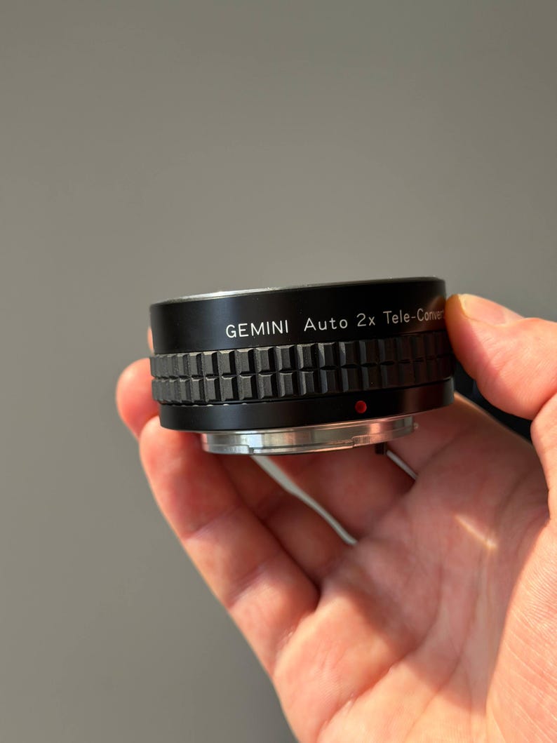 Op de afbeelding: Een zwarte Gemini Auto 2x Tele-Converter lens met een geribbelde grip. De lens heeft zilveren accenten en een rode stip. De tekst "GEMINI Auto 2x Tele-Converter" staat op de lens.