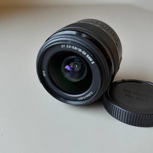 May include: A black camera lens with the text "DT 3.5-5.6/18-55 SAM II" and "0.25m/0.82ft". The lens is partially covered by a black lens cap with the text "TAMRON FOR SONY". The lens is angled slightly to the left.