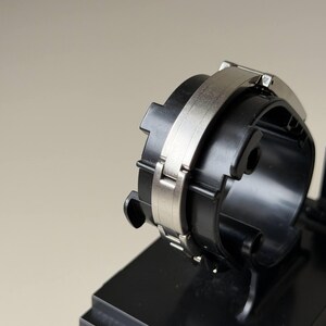 May include: A black and silver watch component. It has a circular design with a silver band and a black base. The watch component is displayed on a black stand against a neutral background.