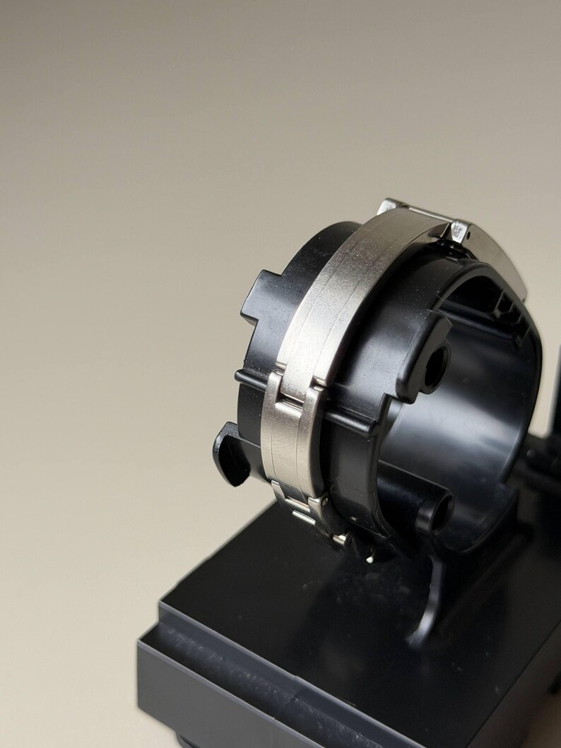 May include: A black and silver watch component. It has a circular design with a silver band and a black base. The watch component is displayed on a black stand against a neutral background.