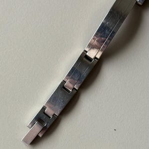May include: A silver wristwatch with a rectangular face and a matching link bracelet. The watch face has a light blue hue. The bracelet is made of flat, rectangular links that connect with a clasp.