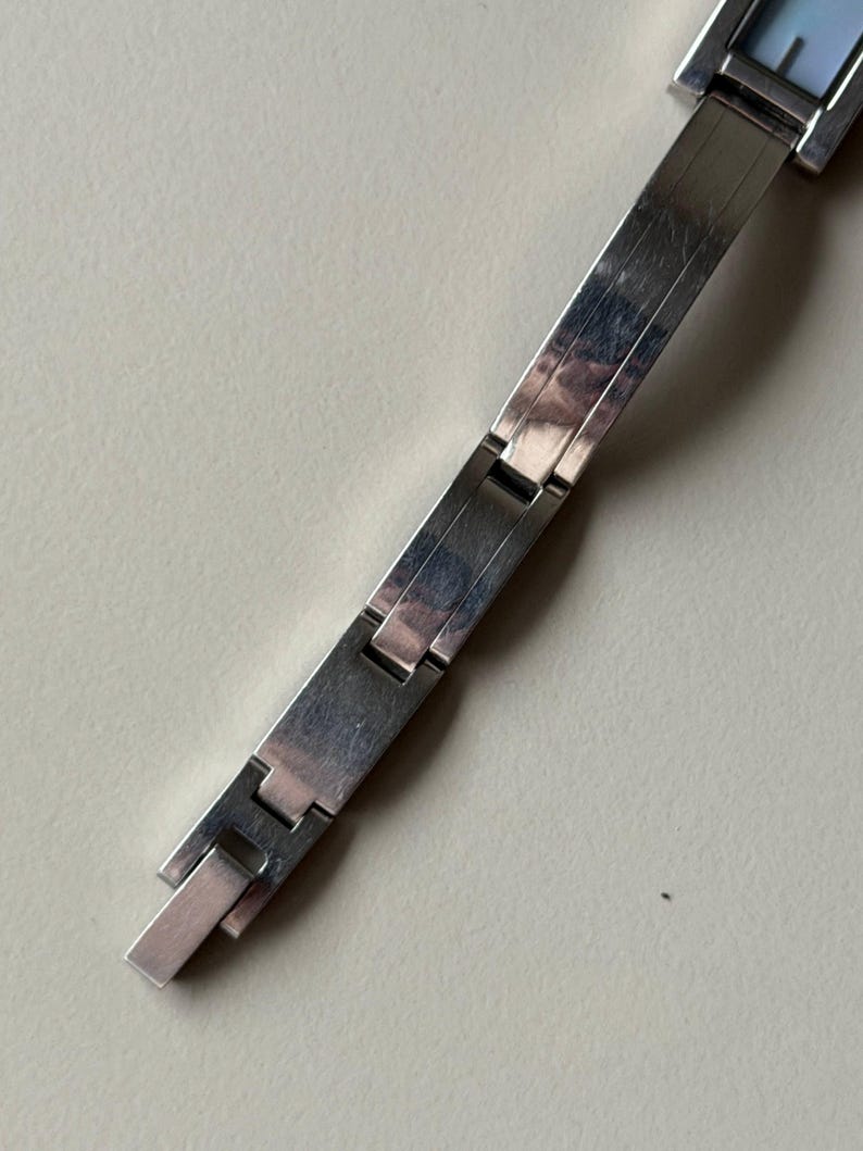 May include: A silver wristwatch with a rectangular face and a matching link bracelet. The watch face has a light blue hue. The bracelet is made of flat, rectangular links that connect with a clasp.