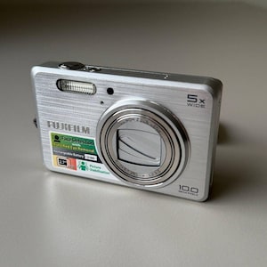 May include: A silver Fujifilm digital camera featuring a 5x wide lens, 10.0 megapixels, and a 2.7-inch LCD screen. The front body displays text including Face Detection, Auto Red Eye Removal, Rechargeable Battery Li-ion, and Picture Stabilization.