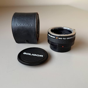 Puede incluir: Un objetivo Soligor MP Auto Tele Converter 2x negro con una tapa de objetivo negra y una funda de cuero negra. La tapa del objetivo y la funda tienen el nombre de la marca Soligor impreso. El objetivo tiene detalles plateados.