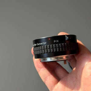Op de afbeelding: Een zwarte teleconverter lensadapter met de tekst "Tele-Converter" en "P/K" aan de zijkant. De adapter heeft een geribbelde grip en een zilverkleurige basis. Een kleine rode stip is zichtbaar aan de bovenkant.