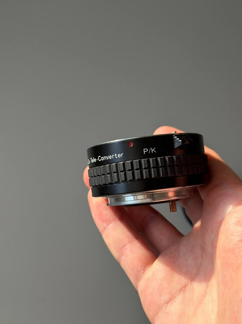 Op de afbeelding: Een zwarte teleconverter lensadapter met de tekst "Tele-Converter" en "P/K" aan de zijkant. De adapter heeft een geribbelde grip en een zilverkleurige basis. Een kleine rode stip is zichtbaar aan de bovenkant.