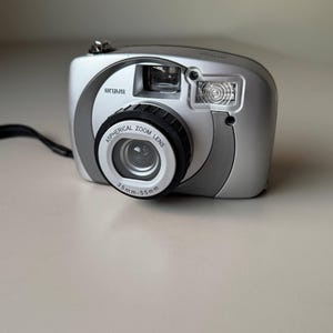 以下が含まれることがあります： 銀色のArgusデジタルカメラ、非球面ズームレンズ付き。レンズは35mm〜55mmです。カメラには内蔵フラッシュと黒いリストストラップが付いています。カメラは明るい色の表面にあります。