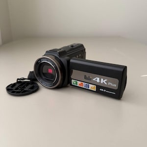 Op de afbeelding: Een zwarte digitale camcorder met een uitklapbaar scherm waarop de tekst 4K Plus en 48.0 Megapixel staat. De lenscilinder bevat de tekst 16X Power Zoom en een losse zwarte ronde lensdop bevindt zich aan de linkerkant van het apparaat.