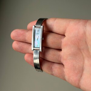 May include: A silver rectangular wristwatch with a mother-of-pearl face. The watch has a slender band and is held in a hand. The watch face displays an iridescent sheen, and the band has a textured design.
