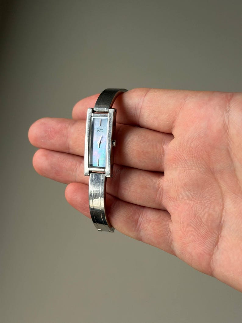 May include: A silver rectangular wristwatch with a mother-of-pearl face. The watch has a slender band and is held in a hand. The watch face displays an iridescent sheen, and the band has a textured design.