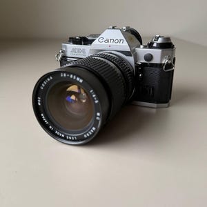 Könnte beinhalten: Eine Canon AE-1 Program 35mm Filmkamera mit einem Cosina 28-85mm Objektiv. Das Kameragehäuse ist silber und schwarz, mit dem Canon-Logo. Das Objektiv ist schwarz mit weißer Schrift, auf einer neutralen Oberfläche.