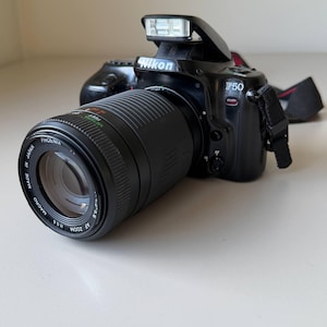 May include: Black Nikon F50 camera with a Phoenix AF Zoom lens. The camera has a built-in flash and a black strap. The lens is marked with measurements in feet and meters.