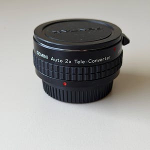 May include: A black Gemini Auto 2x Tele-Converter lens. The lens has a ridged grip and a red dot indicator. The top of the lens has the Gemini logo and text. The lens is designed for photography.