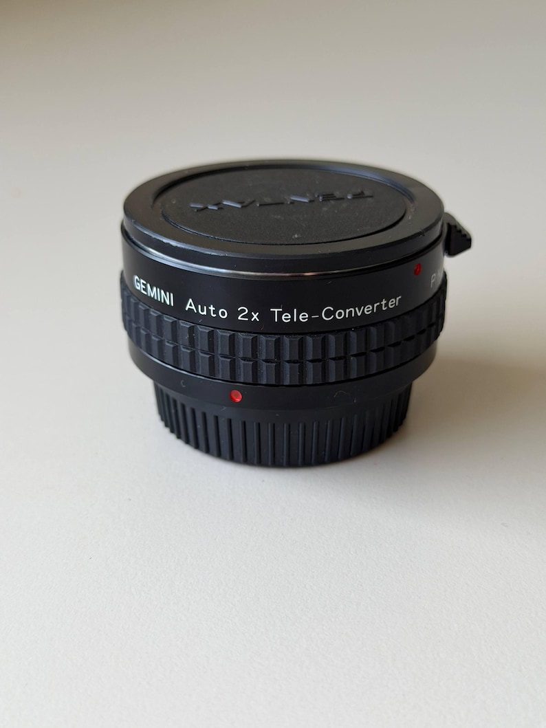 Op de afbeelding: Een zwarte Gemini Auto 2x Tele-Converter lens. De lens heeft een geribbelde grip en een rode puntindicator. De bovenkant van de lens heeft het Gemini-logo en de tekst. De lens is ontworpen voor fotografie.