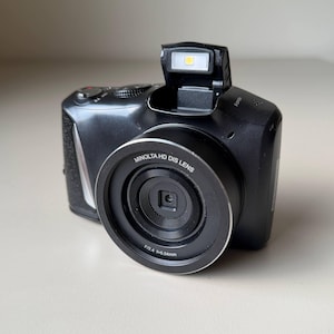 May include: A black Minolta HD DIS lens digital camera with a pop-up flash. The camera has a silver trim around the lens. The lens has the text "MINOLTA HD DIS LENS".