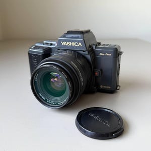 May include: A black Yashica camera with a lens and lens cap. The camera has the word "Yashica" printed in gold on the top and the lens has the measurements 35-70mm 1:3.3-4.5. The lens cap also has the word "Yashica" printed on it.