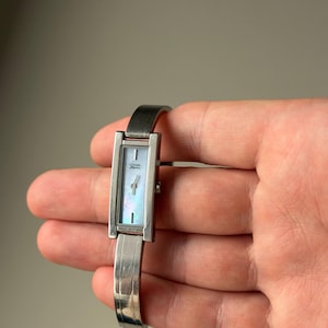 May include: A silver Citizen watch with a rectangular face and a mother-of-pearl dial. The watch has a silver band and a minimalist design. The watch face measures approximately 1.3 cm in width.