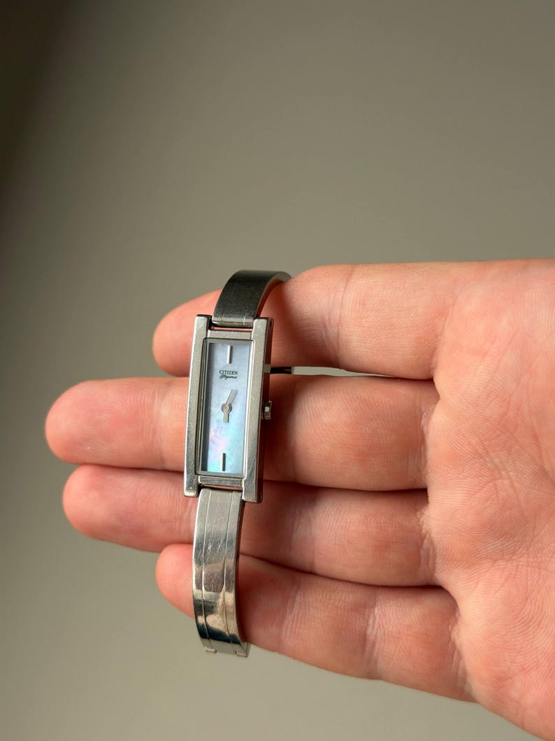 May include: A silver Citizen watch with a rectangular face and a mother-of-pearl dial. The watch has a silver band and a minimalist design. The watch face measures approximately 1.3 cm in width.