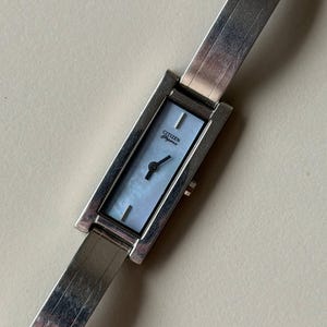 May include: A rectangular Citizen Elegance watch with a silver strap and a light blue face. The watch face has a mother-of-pearl effect. The strap is made of silver links.