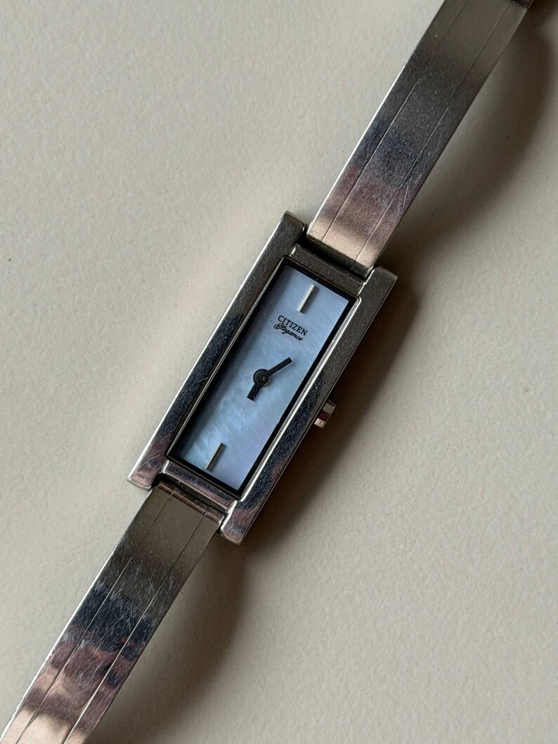 May include: A rectangular Citizen Elegance watch with a silver strap and a light blue face. The watch face has a mother-of-pearl effect. The strap is made of silver links.