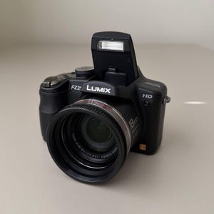 Puede incluir: Una cámara digital negra Panasonic Lumix FZ35 con flash emergente, lente de zoom óptico 18x Vario-Elmarit y marca HD. La cámara cuenta con una empuñadura texturizada, un botón de obturador en la parte superior y un barril de lente con texto.