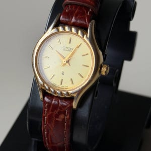May include: A gold-toned Citizen Quartz watch with a cream-colored face and gold hands. The watch has a brown leather band. The watch is displayed on a black stand. The watch face has the text "Citizen Quartz".