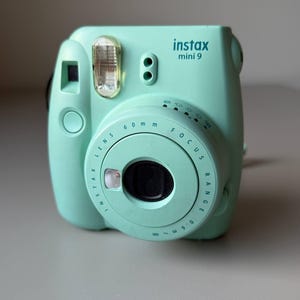 May include: A mint green Instax Mini 9 instant camera. The camera has a built-in flash, a lens with a 60mm focal length, and a focus range dial. The text "instax mini 9" is printed on the top of the camera.