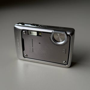 May include: A silver Olympus Stylus SW digital camera with a rectangular body. The front features a lens, a flash, and text reading Olympus, Stylus SW, Shock + Waterproof, 7.1 Megapixel, and Olympus Lens AF Zoom 6.7-20.1mm 1:3.5-5.0.