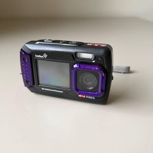 May include: A black and purple Ivation digital camera with a rectangular body, a front-facing screen, and a lens surrounded by a purple frame. The device features the text "ivation", "SHOCKPROOF", and "20 MEGA PIXELS" printed on the front casing.