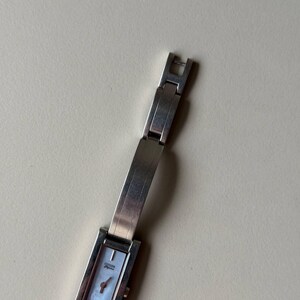 May include: A silver Citizen watch with a rectangular face and a mother-of-pearl dial. The watch has a silver strap and a small, red second hand. The watch is a classic, elegant design.