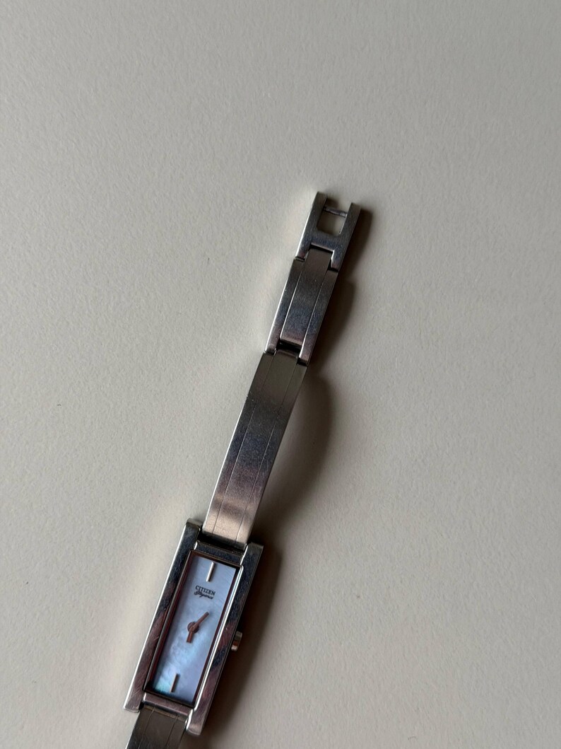 May include: A silver Citizen watch with a rectangular face and a mother-of-pearl dial. The watch has a silver strap and a small, red second hand. The watch is a classic, elegant design.