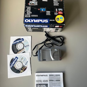 May include: A silver Olympus digital camera with a black strap, a box, two CDs, and a manual. The box is black with the words "Digital camera" and "OLYMPUS" printed on it. The CDs are in white sleeves, and the manual is white with the words "DIGITAL VISION" printed on it.
