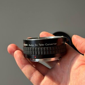 Op de afbeelding: Een zwarte Gemini Auto 2x Tele-Converter lens, vastgehouden in een hand. De lens heeft een geribbeld ontwerp en de tekst "GEMINI Auto 2x Tele-Converter" is op de zijkant gedrukt. Een rode stip is zichtbaar.
