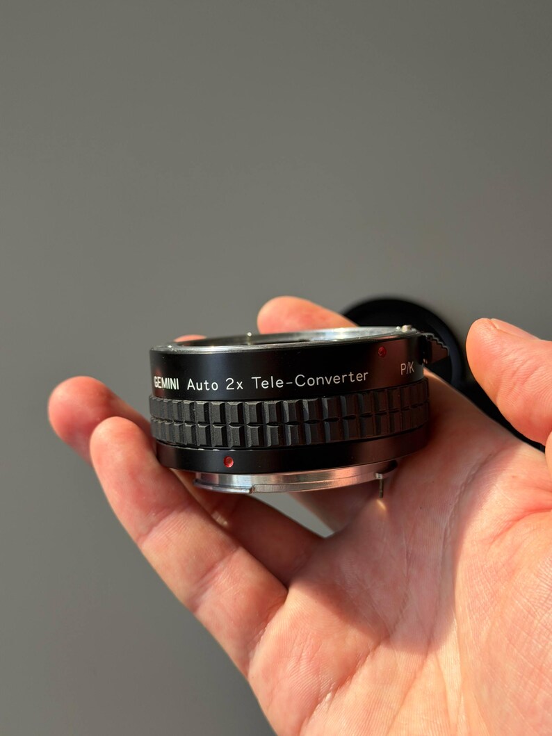 Op de afbeelding: Een zwarte Gemini Auto 2x Tele-Converter lens, vastgehouden in een hand. De lens heeft een geribbeld ontwerp en de tekst "GEMINI Auto 2x Tele-Converter" is op de zijkant gedrukt. Een rode stip is zichtbaar.