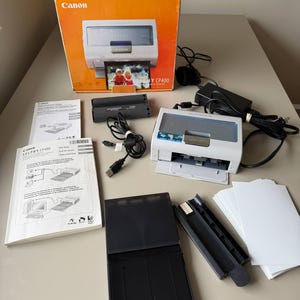 Op de afbeelding: Een Canon Selphy CP400 fotoprinterskit. De kit bevat de printer, voedingsadapter, USB-kabel, papierlade, inktcartridge en handleidingen. De printer is wit en grijs, met een foto van twee personen.