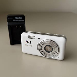 May include: A white Kodak digital camera with a 10 megapixel sensor and a Kodak Retinar Aspheric All Glass Lens, positioned next to a black Kastar battery charger. The camera features a silver lens ring and a small flash unit on the front.
