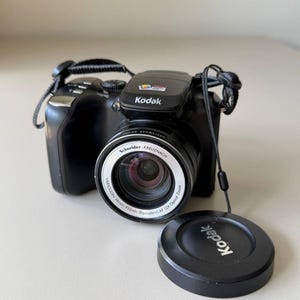 Puede incluir: Una cámara digital Kodak negra con un objetivo Schneider-Kreuznach Variogon de 36mm-432mm, zoom óptico de 12x y estabilizador de imagen. La cámara incluye una correa negra y una tapa de objetivo negra con el logotipo de Kodak impreso en blanco.