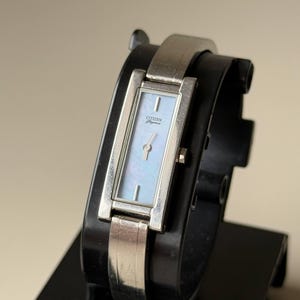May include: A silver Citizen Elegance watch with a rectangular face and a light blue mother-of-pearl dial. The watch has a silver band and is displayed on a black stand. The watch face has the Citizen Elegance logo.