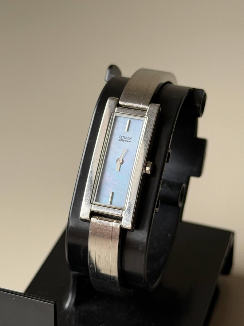 May include: A silver Citizen Elegance watch with a rectangular face and a light blue mother-of-pearl dial. The watch has a silver band and is displayed on a black stand. The watch face has the Citizen Elegance logo.