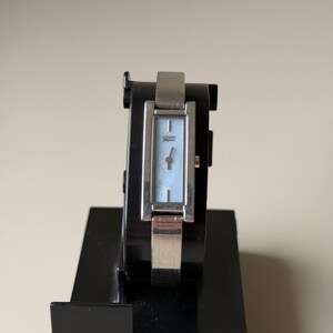 May include: A silver Citizen Elegance watch with a rectangular face and a light blue mother-of-pearl dial. The watch is displayed on a black stand. The watch has a silver band.