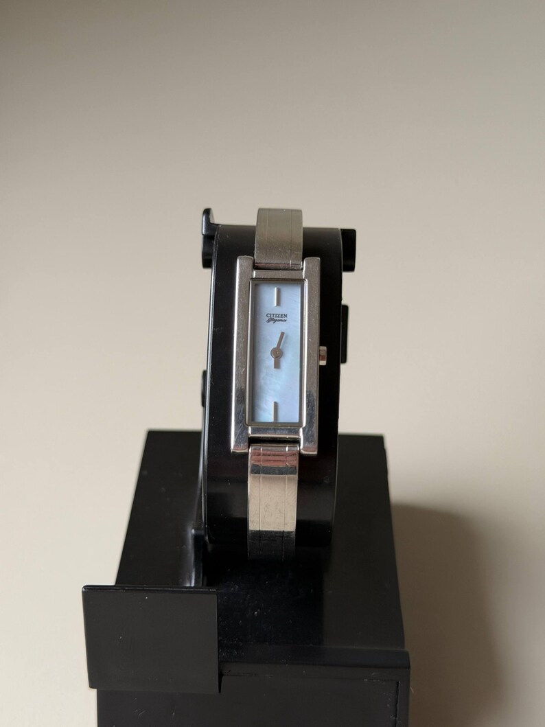May include: A silver Citizen Elegance watch with a rectangular face and a light blue mother-of-pearl dial. The watch is displayed on a black stand. The watch has a silver band.